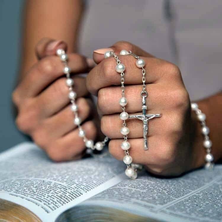 Hands holding a Rosary in prayer over an open Bible